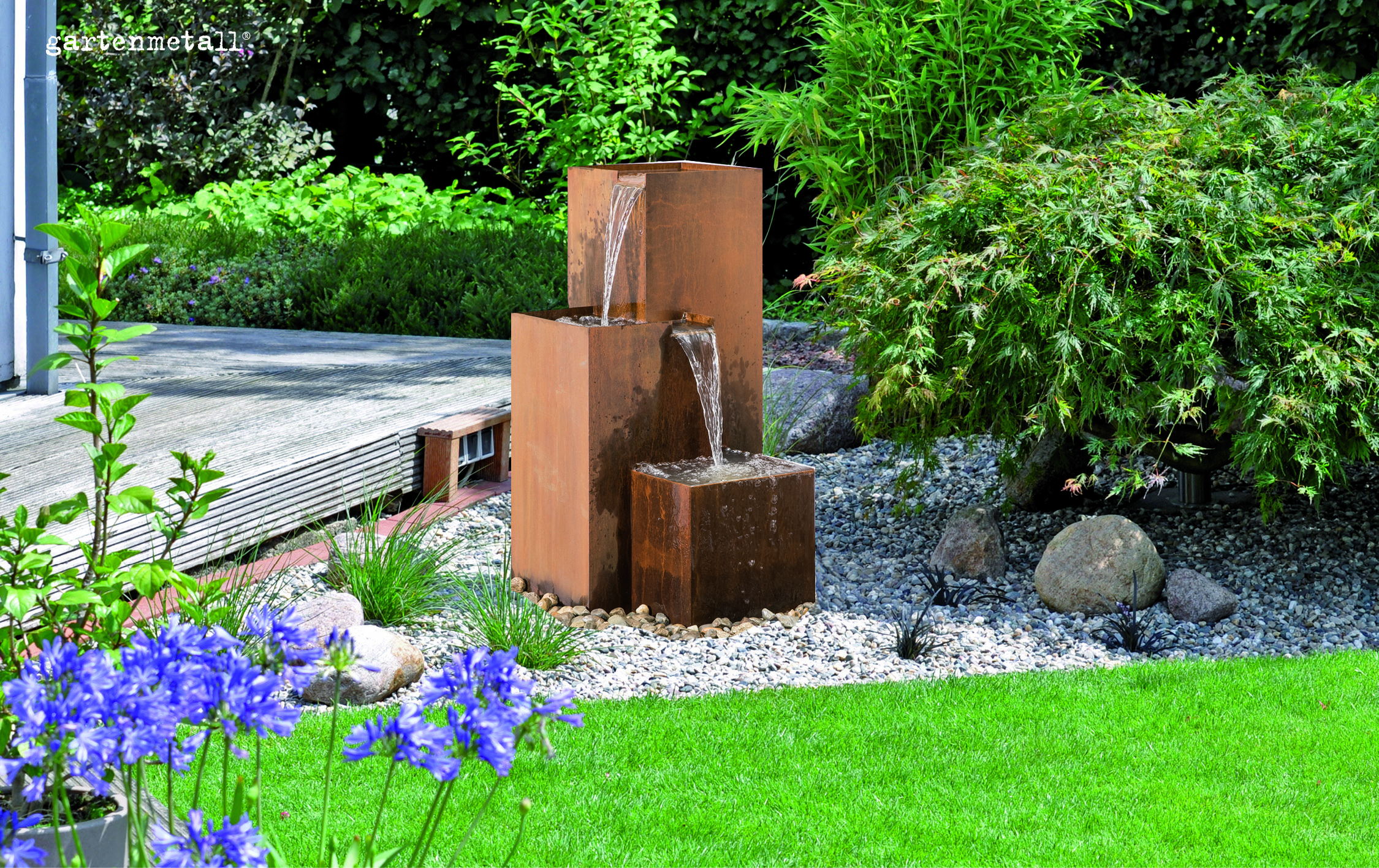 Moderner Brunnen fließt in drei Stufen mit klarem Wasser, umgeben von grünem Garten, Steinen und blühenden Pflanzen. Text: 'gartenmetall'.