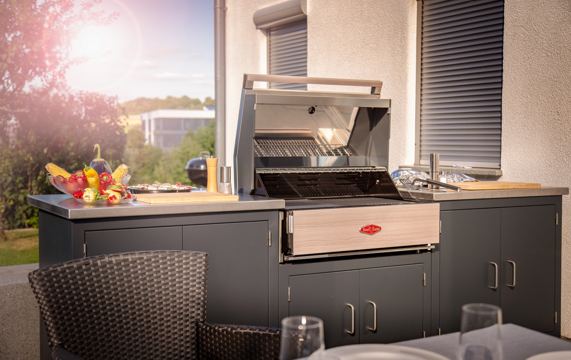 Grill steht geöffnet, ungenutzt, auf einer Außenküche mit Gemüsearrangement. Sonnenlicht und Grünflächen im Hintergrund. Text auf Grill: 'Beef Eater'.