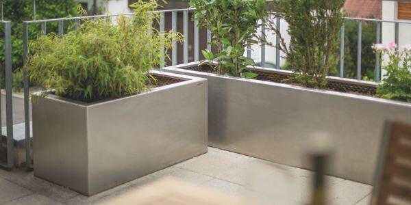 Pflanzkübel aus Metall, bepflanzt mit grünem Bambus und anderen Pflanzen, stehen auf einer Terrasse neben einem Metallgeländer, umgeben von vereinzelten, unscharfen Gebäudeansichten im Hintergrund.