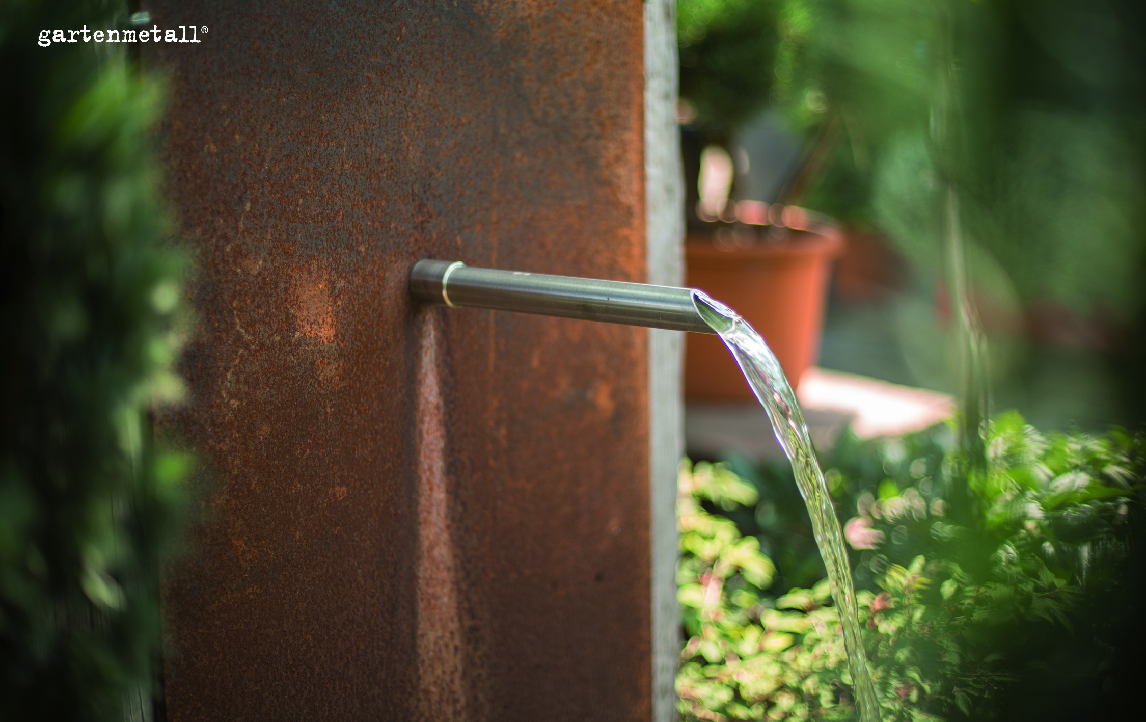 Ein Wasserrohr schüttet einen klaren Wasserstrahl aus an einer rostigen Metallwand, umgeben von grünen Pflanzen im Garten. Text: 'gartenmetall®'.