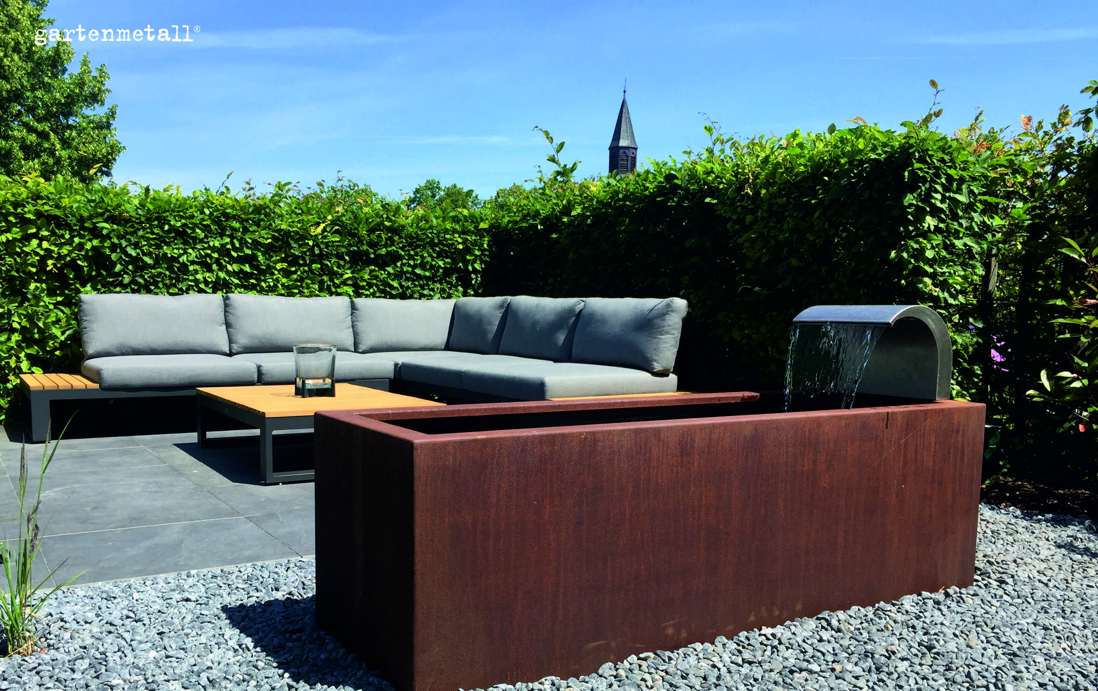 Ein rechteckiger Wasserbrunnen fließt aus Metall in einem Garten, umgeben von Kies. Im Hintergrund steht ein graues Sofa vor einer grünen Hecke. Text: 'gartenmetall®'.