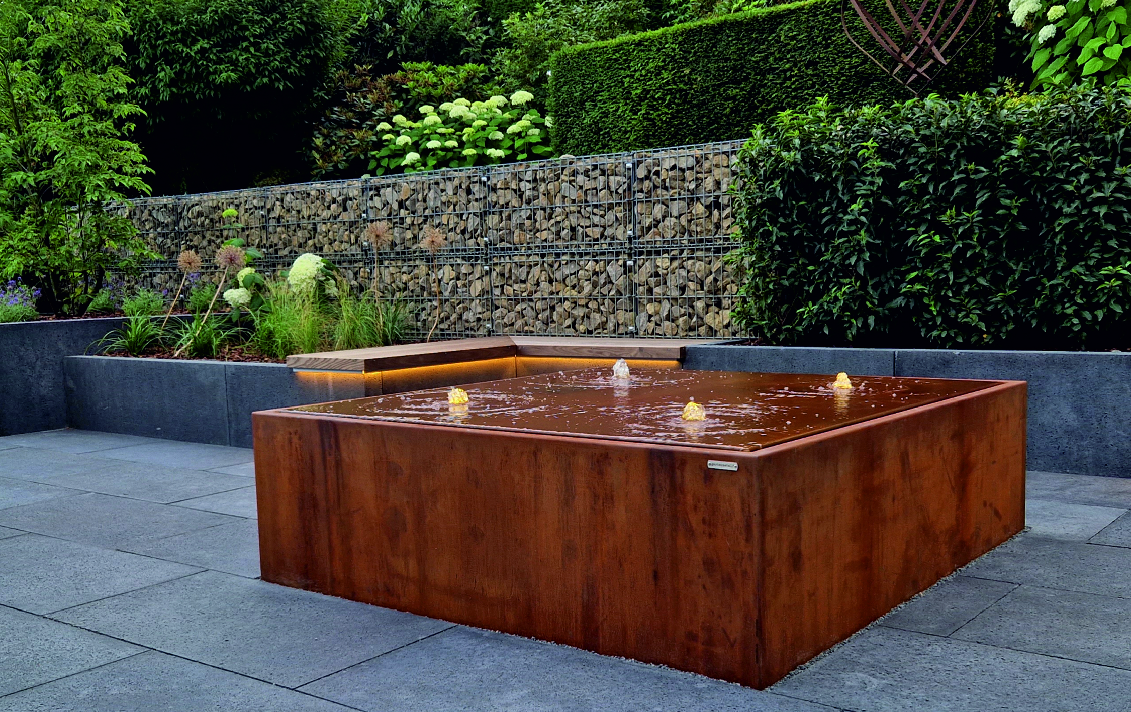 Ein rechteckiger Brunnen mit mehreren Wasserfontänen sprudelt in einem modernen Garten, umgeben von Pflanzen, Gabionenwänden und gepflegten Hecken auf einem gepflasterten Boden.