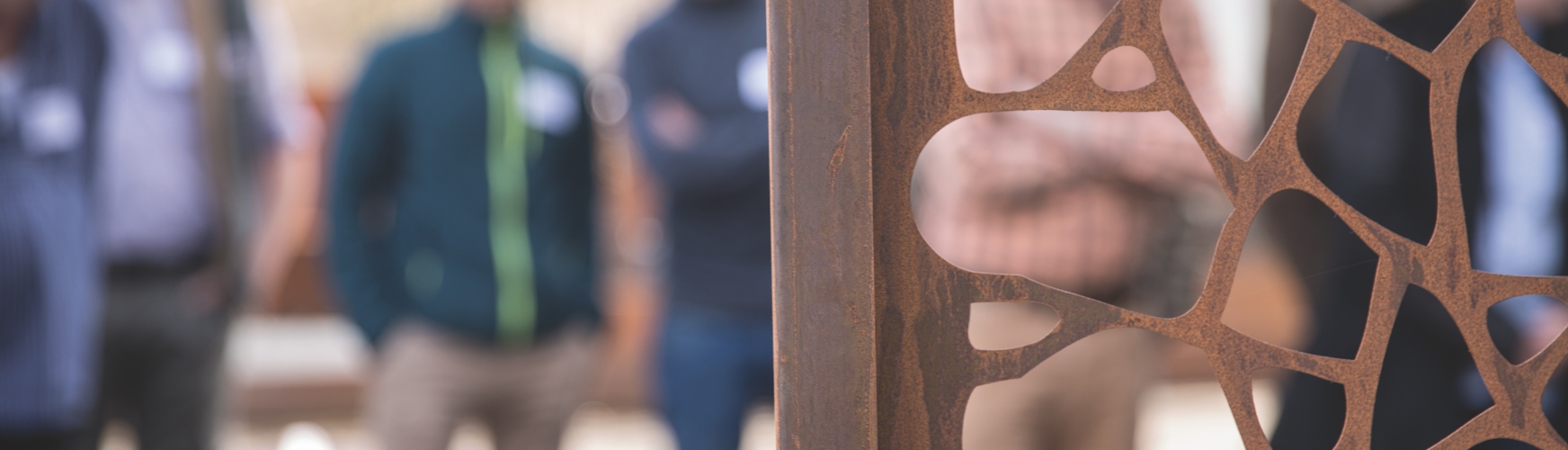 Ein Metallgitter mit abstrakten Mustern steht im Vordergrund, während im Hintergrund unscharfe Menschen stehen und diskutieren. Die Umgebung wirkt wie ein offener, urbaner Raum.