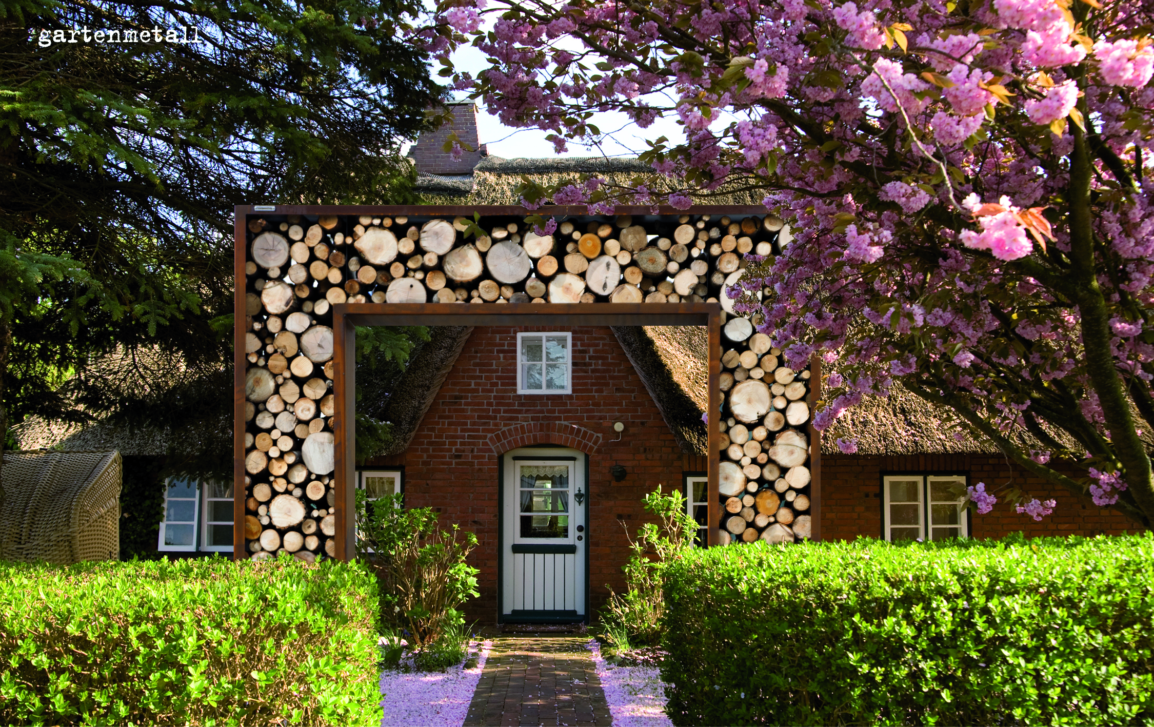 Holzverzierte Torbogen, rahmt Eingangsbereich eines roten Backsteinhauses. Umgeben von blühenden Kirschblütenbäumen und gepflegten Hecken. Text im Bild: 'gartenmetall'.
