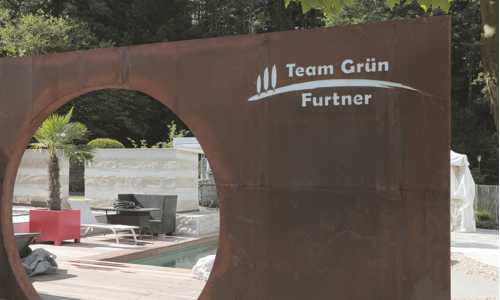 Metallische Skulptur mit kreisförmiger Aussparung, beschriftet mit „Team Grün Furtner“, steht in einem modernen Gartenbereich mit Palmen und Sitzmöbeln.