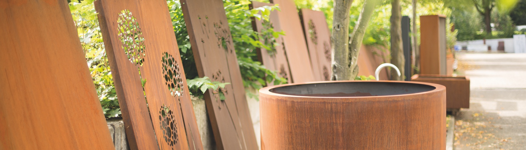 Metalltafeln mit floralen Ausstanzungen lehnen an einer Wand in einem grünen, sonnigen Außenbereich. Im Vordergrund steht ein großer, rostfarbener Pflanzkübel neben einem Weg.