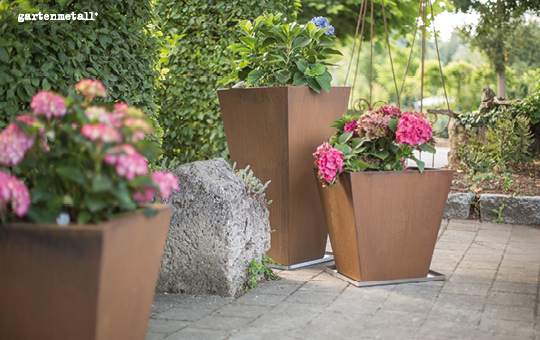Pflanzkübel stehen auf gepflastertem Boden, gefüllt mit blühenden Hortensien. Im Hintergrund sind Hecken und Bäume sichtbar. Oben links steht der Text 'gartenmetall'.