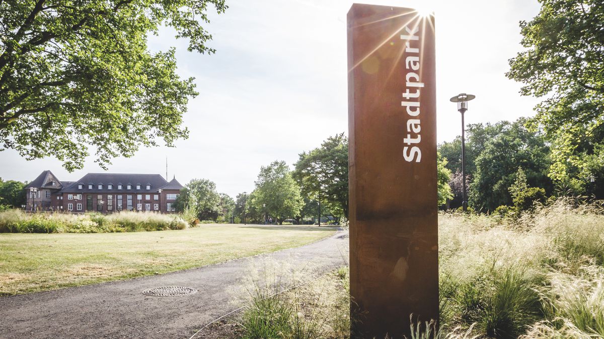 Säule mit Text 'Stadtpark' steht am Weg, umgeben von grünem Rasen und Bäumen, während ein rotes Backsteingebäude im Hintergrund sichtbar ist.