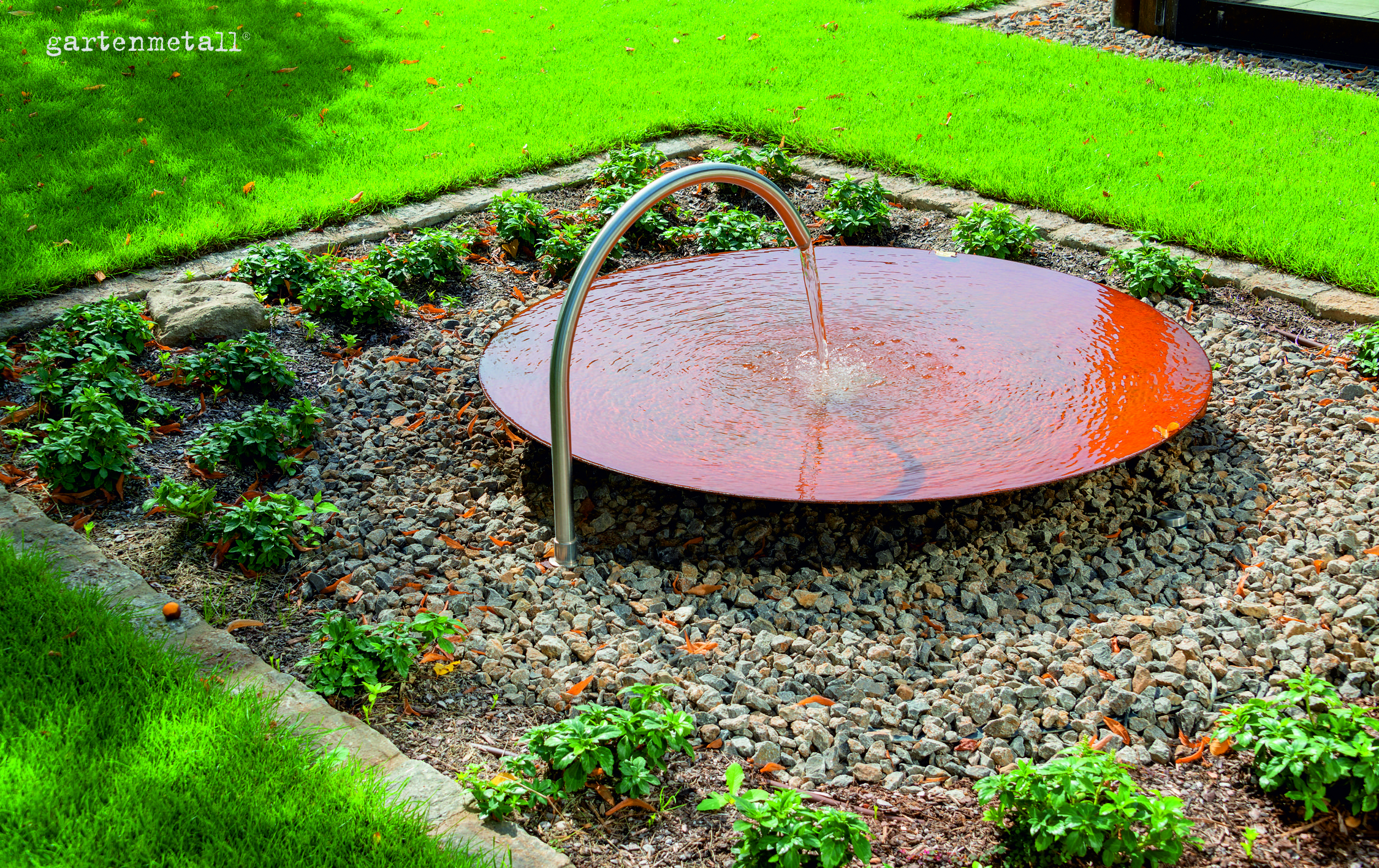 Ein runder, flacher Wasserauslauf mit Edelstahlbogen lässt Wasser auf eine rote Metallschale in einem steinernen Beet plätschern. Umgeben von grünem Rasen und kleinen Pflanzen. Text: gartenmetall®.