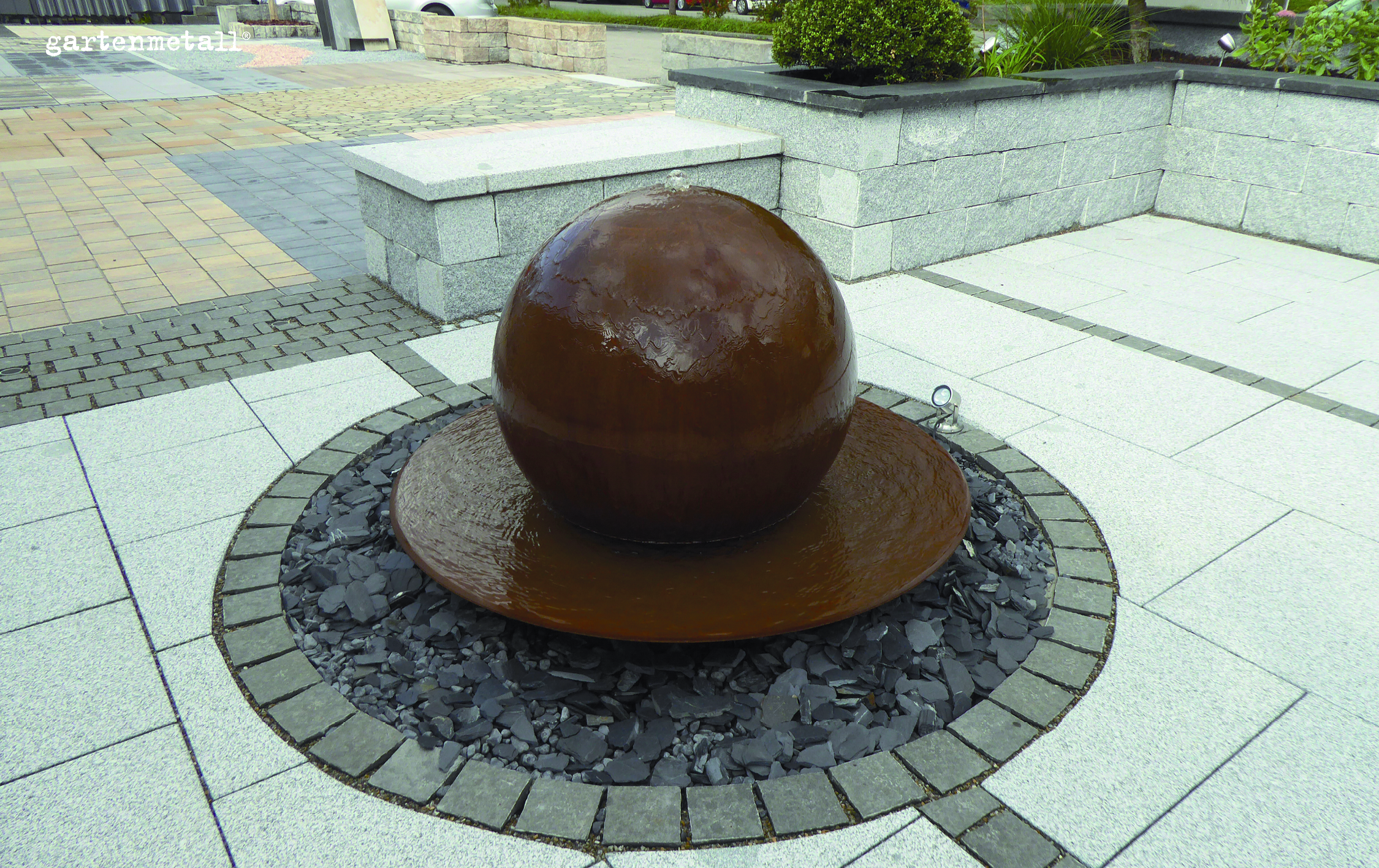 Kugelbrunnen dreht sich auf einer Wasserfläche, umgeben von grauem Pflastersteinen und Steinplatten in einer offenen, gepflasterten Anlage. Text: 'gartenmetall®'.