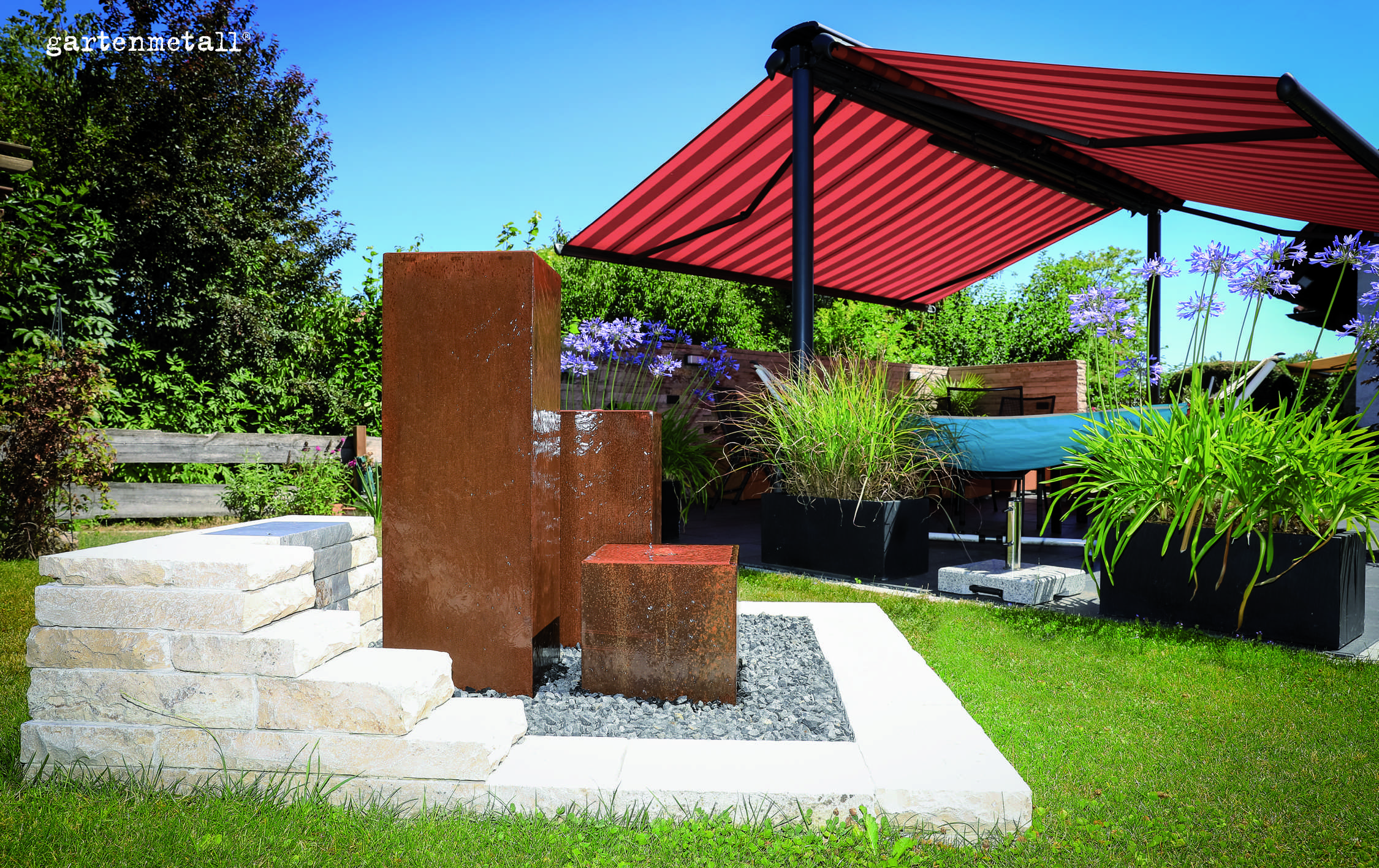 Rostfarbene Metallskulpturen sprudeln Wasser, umgeben von Steinplatten und Kies, in einem Garten mit üppiger Vegetation und einem roten Sonnendach. Text: gartenmetall.
